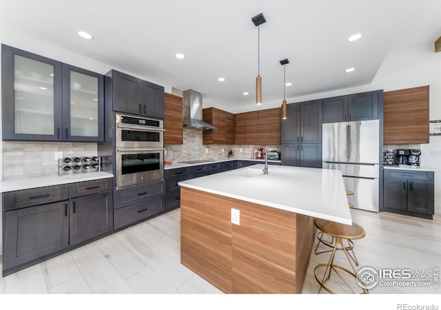 kitchen featuring sink, an island with sink, pendant lighting, stainless steel appliances, and wall chimney range hood