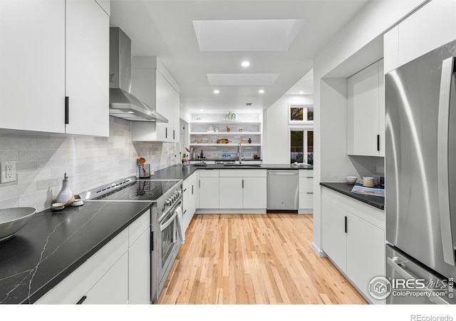 kitchen with sink, a skylight, appliances with stainless steel finishes, wall chimney range hood, and white cabinets