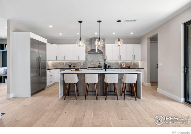 kitchen with a breakfast bar, decorative light fixtures, white cabinetry, stainless steel built in refrigerator, and wall chimney range hood