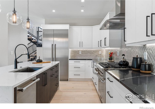 kitchen with sink, dark stone countertops, high quality appliances, wall chimney range hood, and white cabinets