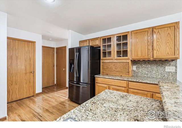 kitchen with tasteful backsplash, glass insert cabinets, light stone counters, light wood-style floors, and stainless steel refrigerator with ice dispenser