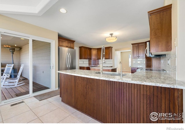 kitchen with light tile patterned flooring, stainless steel refrigerator, backsplash, kitchen peninsula, and light stone countertops