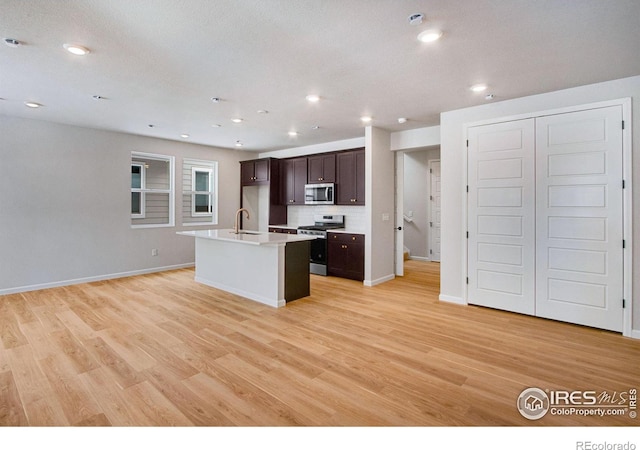 kitchen with sink, a kitchen island with sink, dark brown cabinetry, stainless steel appliances, and light hardwood / wood-style flooring