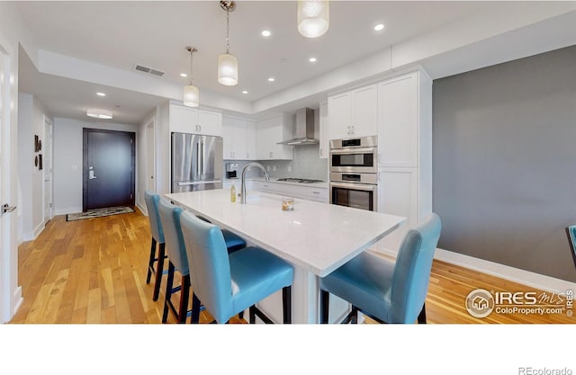 kitchen featuring white cabinetry, an island with sink, hanging light fixtures, stainless steel appliances, and wall chimney range hood