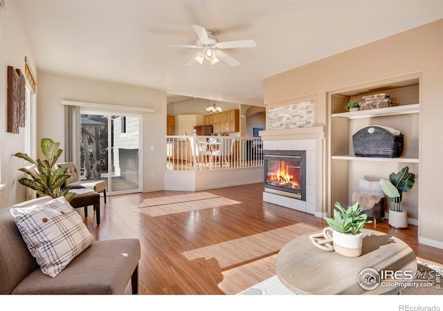 living room featuring a tile fireplace, light wood-style flooring, ceiling fan with notable chandelier, baseboards, and built in features