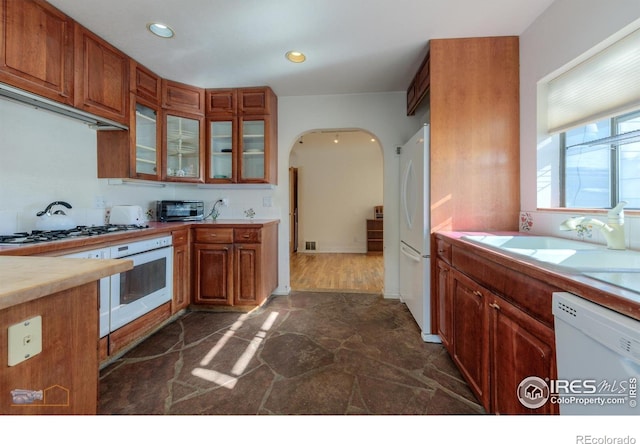 kitchen featuring a sink, brown cabinets, white appliances, arched walkways, and stone finish floor
