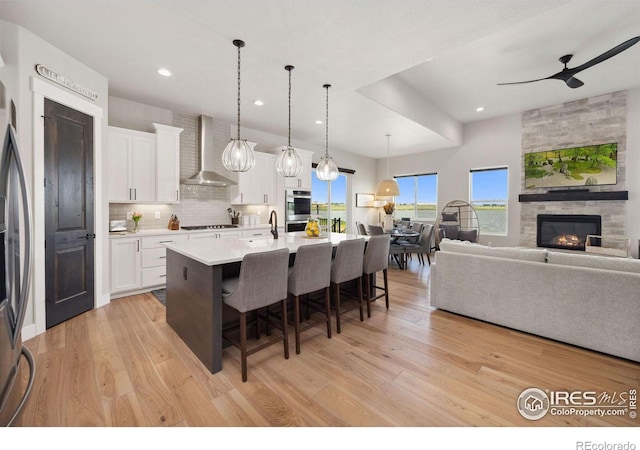 kitchen featuring pendant lighting, a kitchen breakfast bar, white cabinets, a center island with sink, and wall chimney exhaust hood