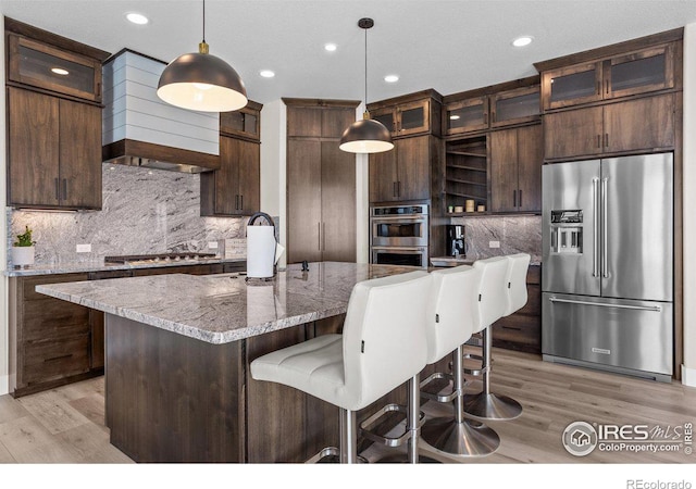 kitchen with pendant lighting, a kitchen island with sink, stainless steel appliances, dark brown cabinetry, and light stone countertops