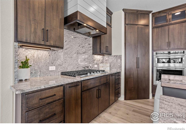 kitchen featuring dark brown cabinets, stainless steel appliances, tasteful backsplash, light stone countertops, and custom exhaust hood