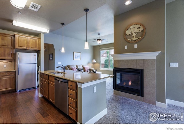 kitchen featuring appliances with stainless steel finishes, a fireplace, an island with sink, sink, and dark wood-type flooring