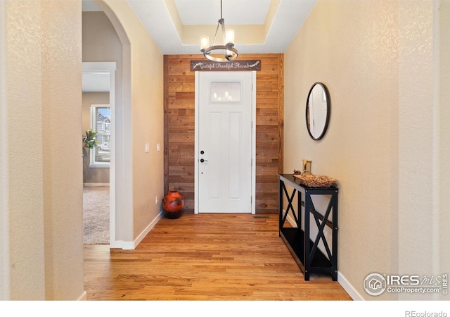 entryway featuring arched walkways, wooden walls, baseboards, light wood finished floors, and a tray ceiling