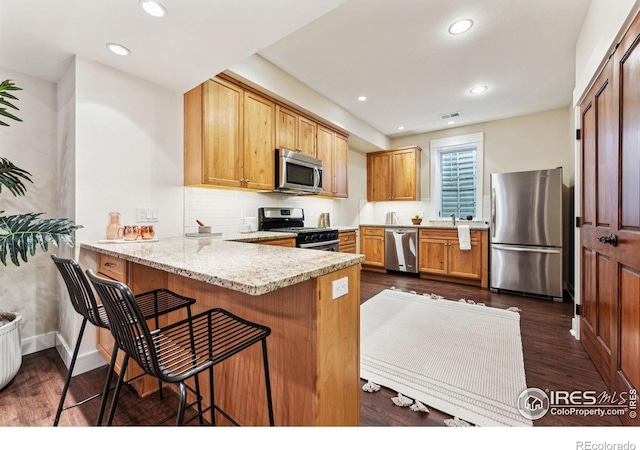 kitchen featuring dark wood-type flooring, a kitchen bar, light stone counters, appliances with stainless steel finishes, and kitchen peninsula