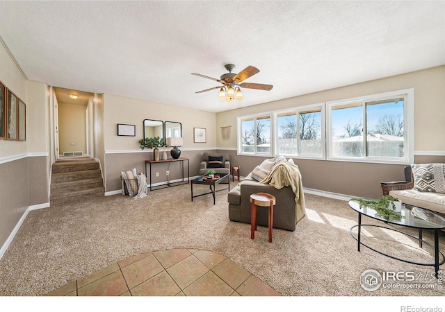 tiled living area featuring a textured ceiling, ceiling fan, carpet floors, visible vents, and stairs