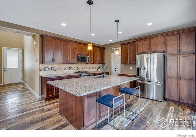kitchen featuring appliances with stainless steel finishes, light stone counters, decorative light fixtures, a kitchen island with sink, and a sink