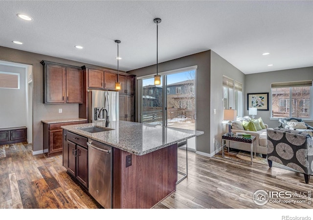 kitchen with a kitchen island with sink, stainless steel appliances, a sink, hanging light fixtures, and light stone countertops