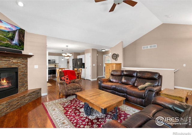 living room featuring vaulted ceiling, a stone fireplace, dark wood-type flooring, and ceiling fan with notable chandelier