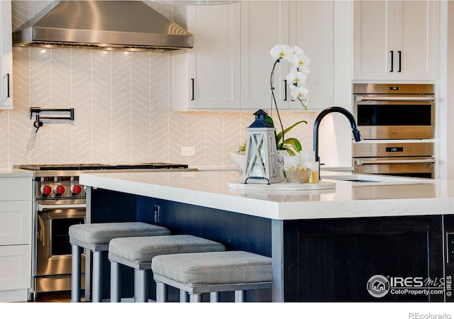 kitchen with appliances with stainless steel finishes, white cabinets, wall chimney range hood, decorative backsplash, and a breakfast bar