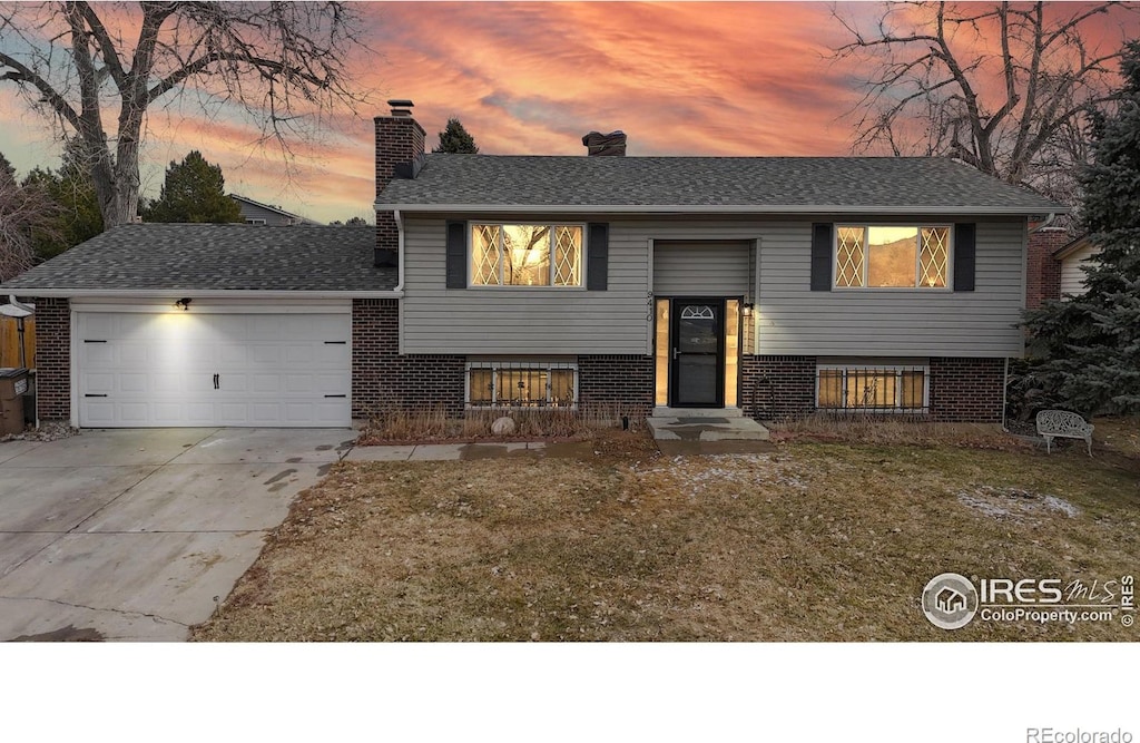 raised ranch with a garage, a chimney, concrete driveway, and brick siding