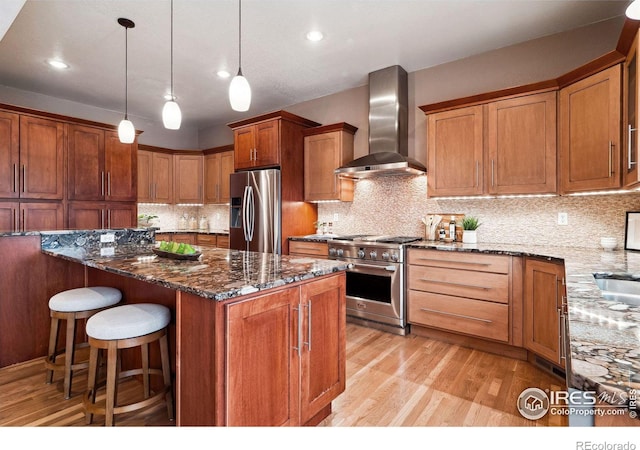 kitchen with light hardwood / wood-style floors, dark stone counters, appliances with stainless steel finishes, wall chimney range hood, and pendant lighting