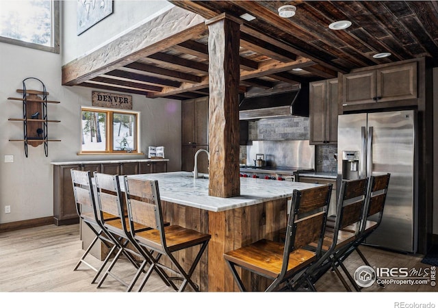 kitchen with an island with sink, custom range hood, decorative backsplash, light hardwood / wood-style floors, and stainless steel fridge with ice dispenser