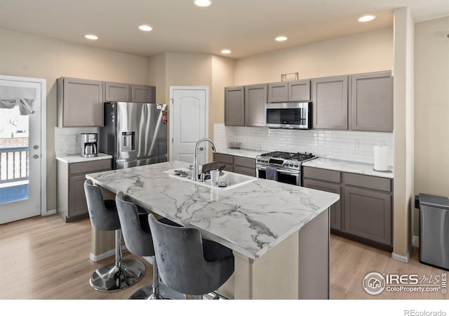 kitchen with stainless steel appliances, a kitchen island with sink, gray cabinetry, and light stone countertops