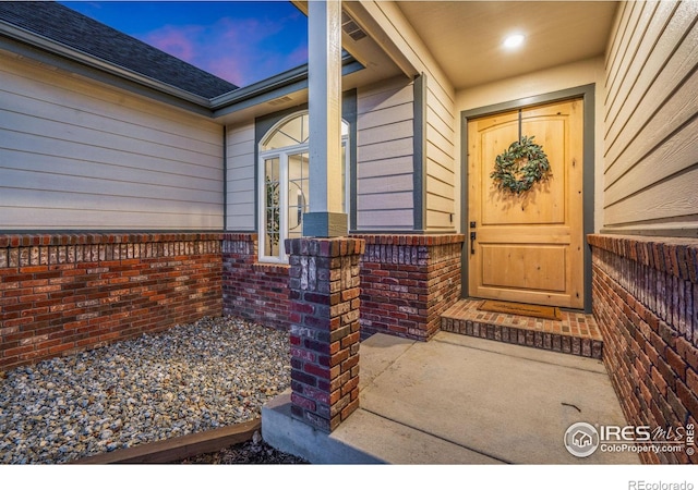 exterior entry at dusk featuring roof with shingles, fence, and brick siding