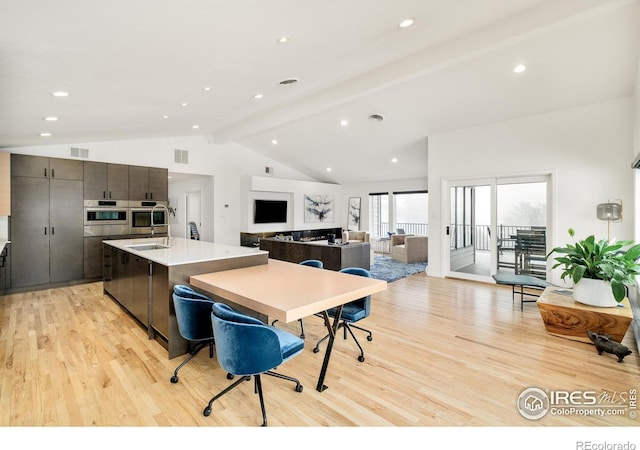 kitchen with stainless steel oven, light wood-style flooring, visible vents, a kitchen island with sink, and vaulted ceiling with beams