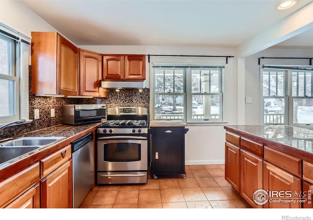 kitchen with under cabinet range hood, appliances with stainless steel finishes, tile counters, tasteful backsplash, and brown cabinetry