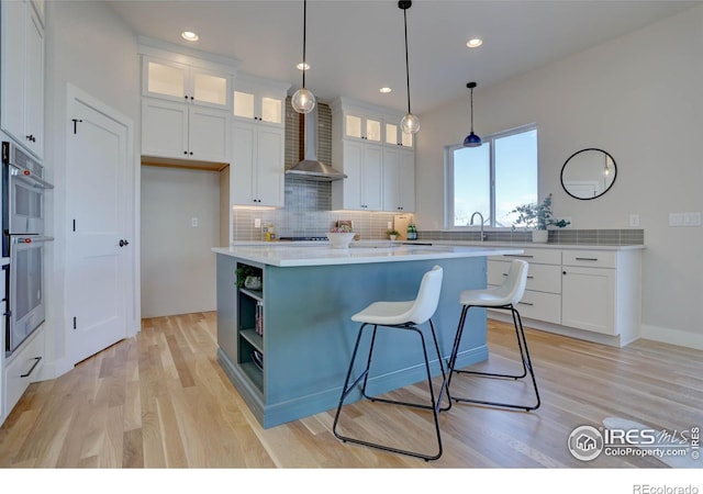 kitchen featuring white cabinets, glass insert cabinets, a center island, light countertops, and wall chimney range hood