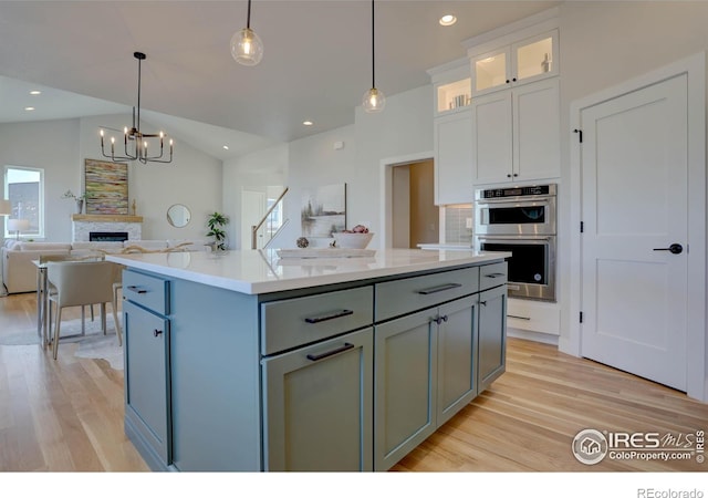 kitchen featuring stainless steel double oven, white cabinets, hanging light fixtures, and an island with sink