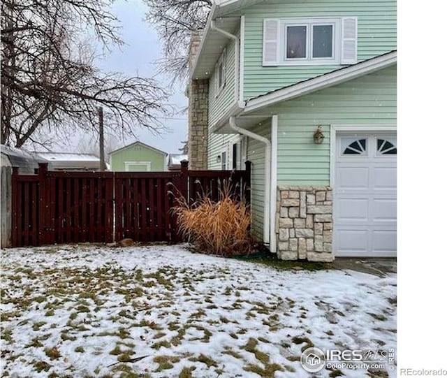 snow covered property with a garage, stone siding, and fence