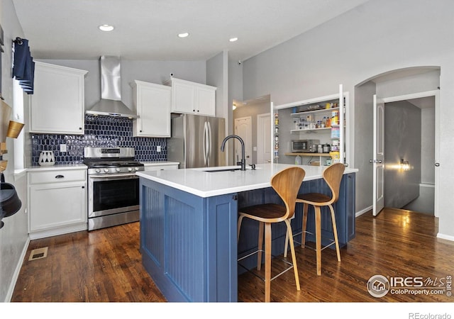 kitchen featuring stainless steel appliances, light countertops, a kitchen island with sink, a sink, and wall chimney range hood