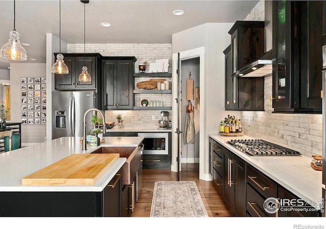 kitchen featuring a kitchen island with sink, dark wood-style floors, appliances with stainless steel finishes, backsplash, and decorative light fixtures
