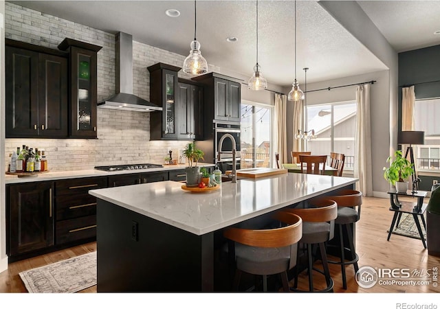 kitchen with appliances with stainless steel finishes, an island with sink, wall chimney exhaust hood, decorative backsplash, and glass insert cabinets