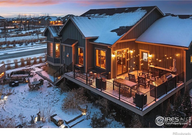 snow covered back of property with board and batten siding, a deck, roof with shingles, and an outdoor fire pit