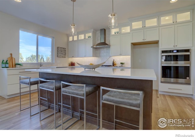 kitchen featuring a kitchen island, glass insert cabinets, light countertops, wall chimney range hood, and double oven