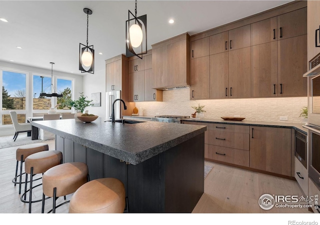 kitchen featuring a center island with sink, custom exhaust hood, dark countertops, hanging light fixtures, and a sink