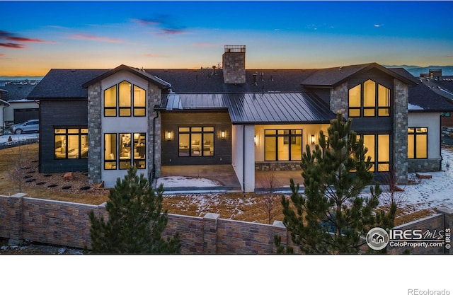 snow covered house with metal roof, a patio area, stone siding, a standing seam roof, and a chimney