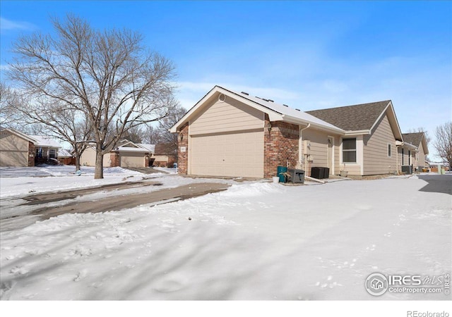 snow covered property with an attached garage, central AC unit, and brick siding