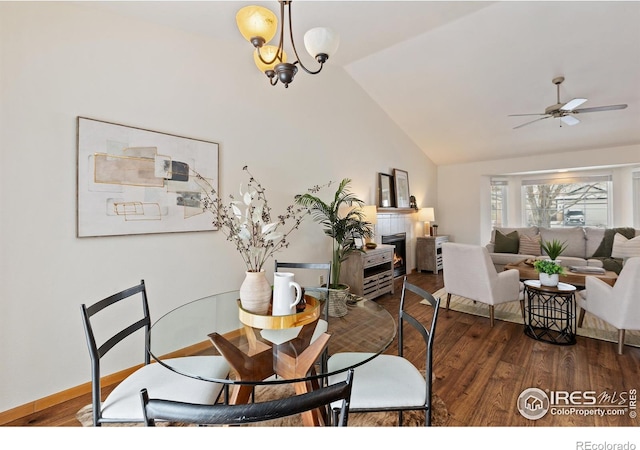 dining space featuring lofted ceiling, ceiling fan with notable chandelier, dark wood-type flooring, a fireplace, and baseboards