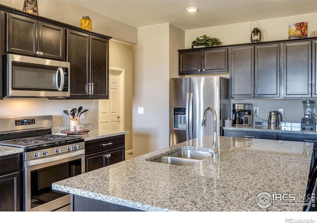 kitchen featuring an island with sink, light stone counters, stainless steel appliances, dark brown cabinets, and a sink