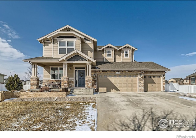 view of front of home featuring roof with shingles, covered porch, concrete driveway, fence, and stone siding