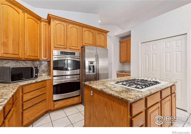 kitchen with light tile patterned floors, light stone countertops, stainless steel appliances, backsplash, and brown cabinets