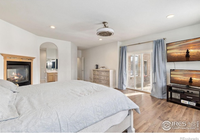 bedroom featuring arched walkways, recessed lighting, ensuite bath, light wood-style floors, and a glass covered fireplace