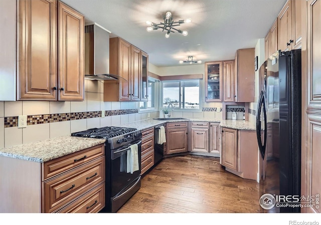 kitchen with glass insert cabinets, a sink, wall chimney range hood, light stone countertops, and black appliances