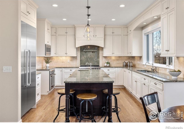 kitchen with an island with sink, dark stone countertops, built in appliances, hanging light fixtures, and a sink