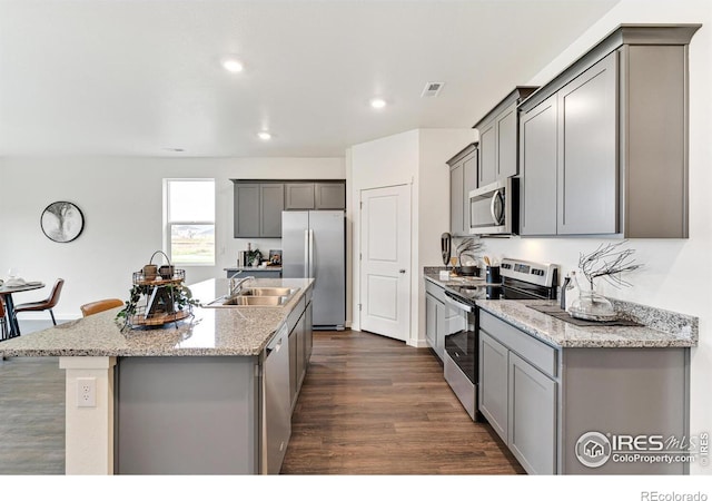 kitchen with a kitchen island with sink, stainless steel appliances, a sink, gray cabinets, and light stone countertops