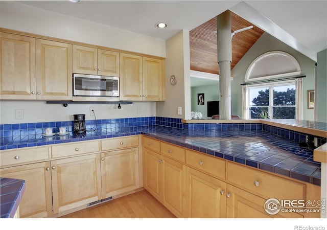 kitchen featuring tile counters, stainless steel microwave, visible vents, and light brown cabinetry