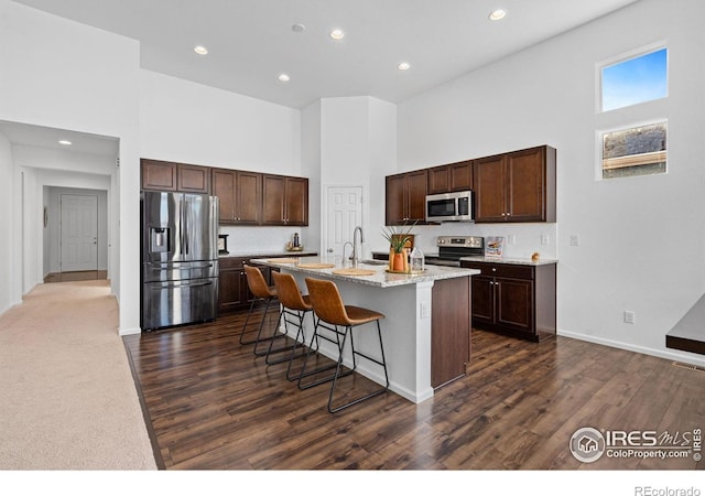 kitchen with stainless steel appliances, a towering ceiling, light stone countertops, an island with sink, and a kitchen bar