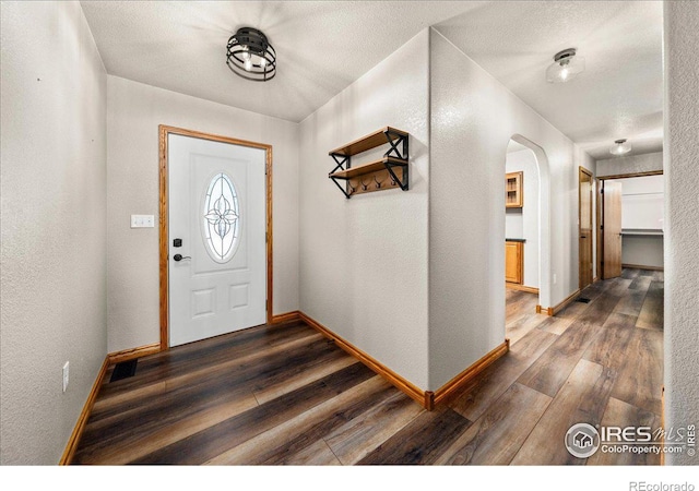 foyer featuring a textured ceiling, a textured wall, arched walkways, baseboards, and dark wood-style floors